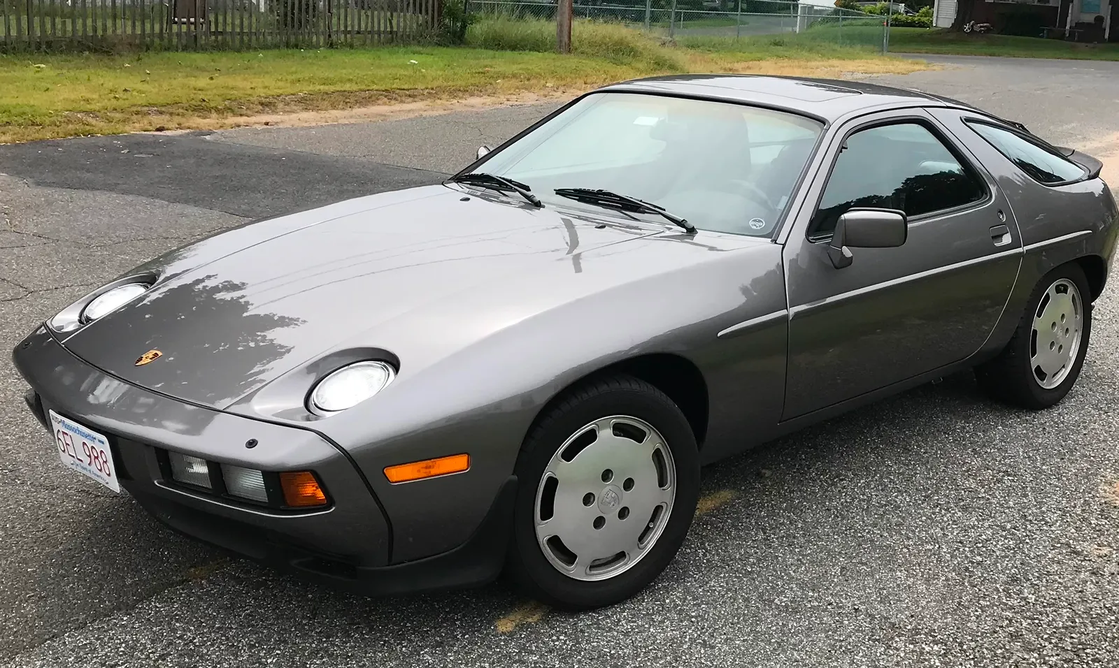 1989 Porsche 928 parked in a parking lot next to a house, surrounded by grass, plants, trees, and a fence