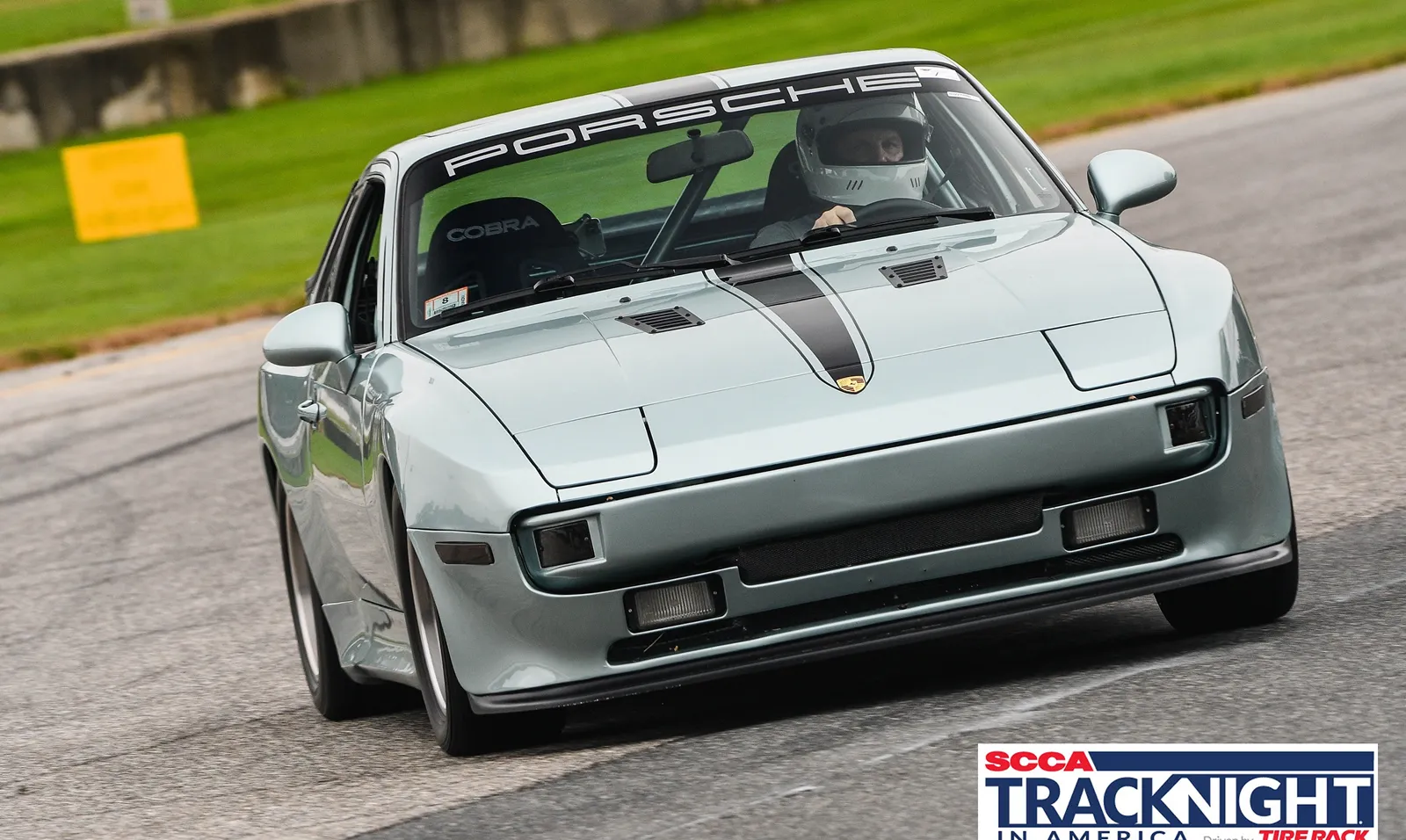 Person wearing a helmet driving a silver Porsche 930 on a race track, surrounded by grass